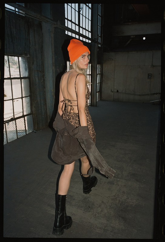Woman in animal-print dress and orange beanie stands in an industrial location, looking back over shoulder with natural light.