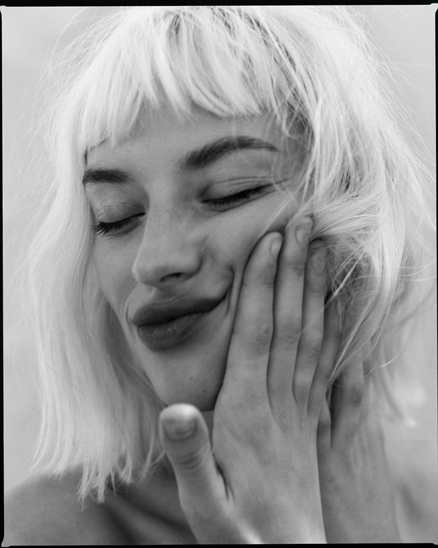 Close-up black and white portrait of a blonde woman with closed eyes and a slight smile, her hand gently touching her cheek.