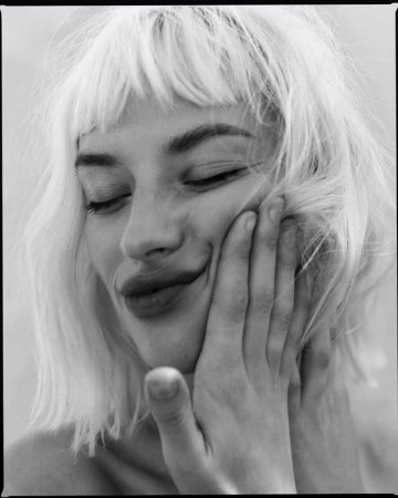 Close-up black and white portrait of a blonde woman with closed eyes and a slight smile, her hand gently touching her cheek.