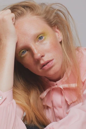 Close-up portrait of an adult female with fair skin, light hair, and yellow eyeshadow, wearing a pink pleated blouse and looking at the camera.