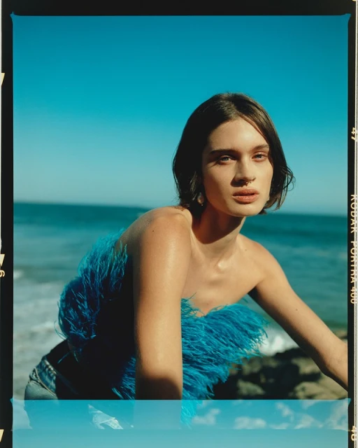 Young adult woman poses in a blue feathered top and jeans against a backdrop of the ocean and sky on a sunny day.
