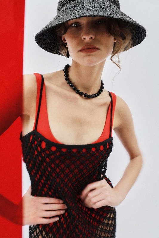 Fashion portrait of a woman wearing a straw hat, black necklace, and a red and black dress against a white background with a red accent.