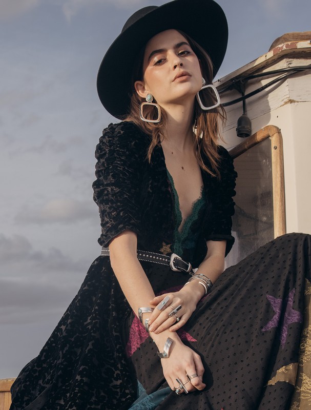 A woman in a black hat and dress poses outdoors, showcasing large silver earrings and rings, with a muted sky in the background.