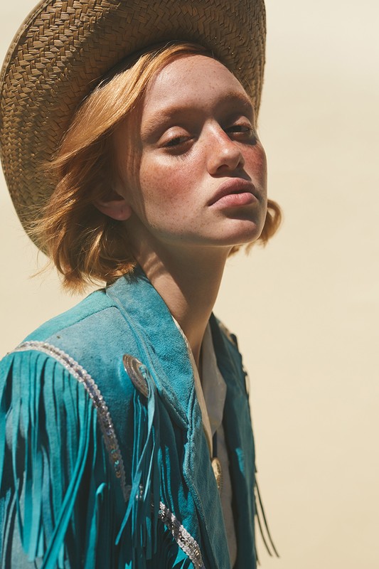 Portrait of a woman with freckles wearing a straw cowboy hat and a fringe turquoise jacket against a neutral-toned background in daylight.