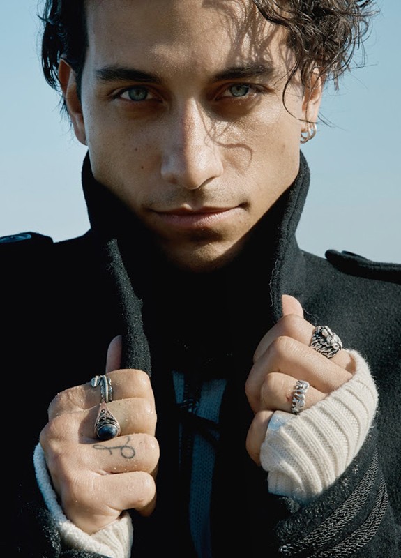 Close-up portrait of a man in a black coat holding the collar, with rings on his fingers and styled curly hair against a clear blue sky