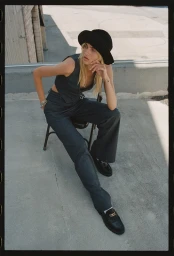 Model in dark vest and trousers wearing black hat and loafers sitting outdoors on chair