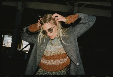 Woman in grey plaid blazer and striped sweater adjusting her blonde hair with sunglasses on indoors