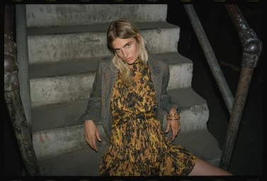 Woman with blonde hair wearing a patterned dress and plaid blazer seated on concrete stairs with metal railings.