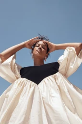 Model in voluminous white dress with black bodice posing with hands on head against clear blue sky in Los Angeles.