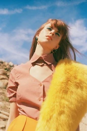 A red-haired woman wearing a pink patterned collared shirt and yellow fur coat outdoors against a bright blue sky.