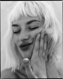 Close-up monochrome portrait of a woman with short blonde hair gently pressing her hand against her cheek.