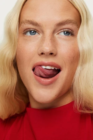 Close-up portrait of a woman with blonde wavy hair and blue eyes wearing a bright red top and licking her lips