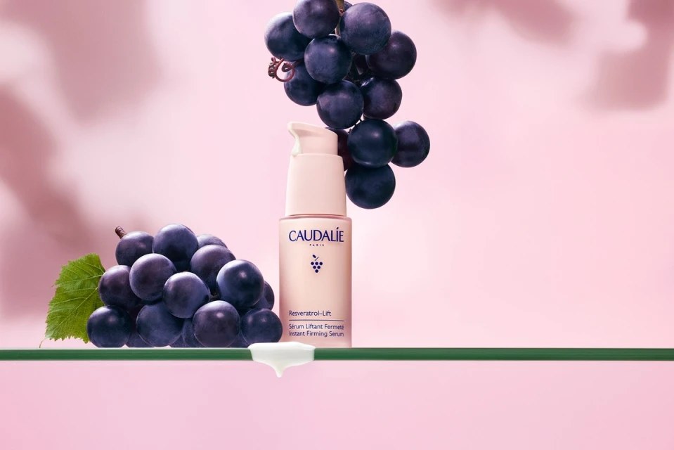 Caudalie Resveratrol Lift serum bottle displayed with clusters of dark purple grapes against a pink backdrop on a glass surface.