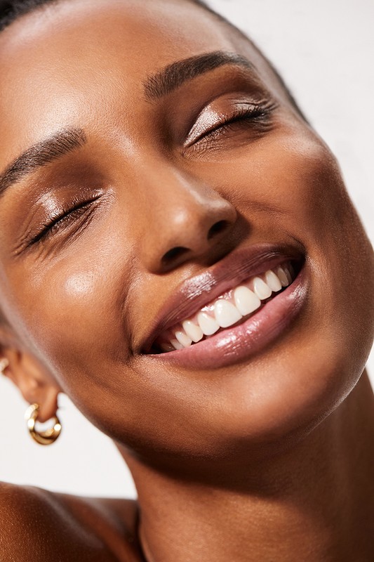 Close-up of a woman with tanned skin and closed eyes, showcasing her radiant complexion and glossy lips, wearing gold hoop earrings.