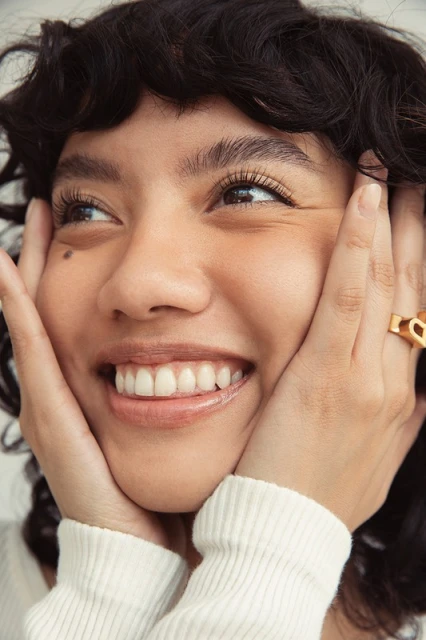 Close-up of a smiling adult female with her hands on her face, showcasing her curly dark hair and gold ring, against a neutral background.