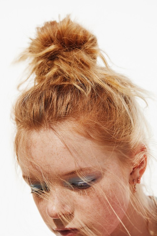 Close-up portrait of a young adult female with fair skin and freckles, with red hair styled in a messy bun, set against a plain white backdrop.