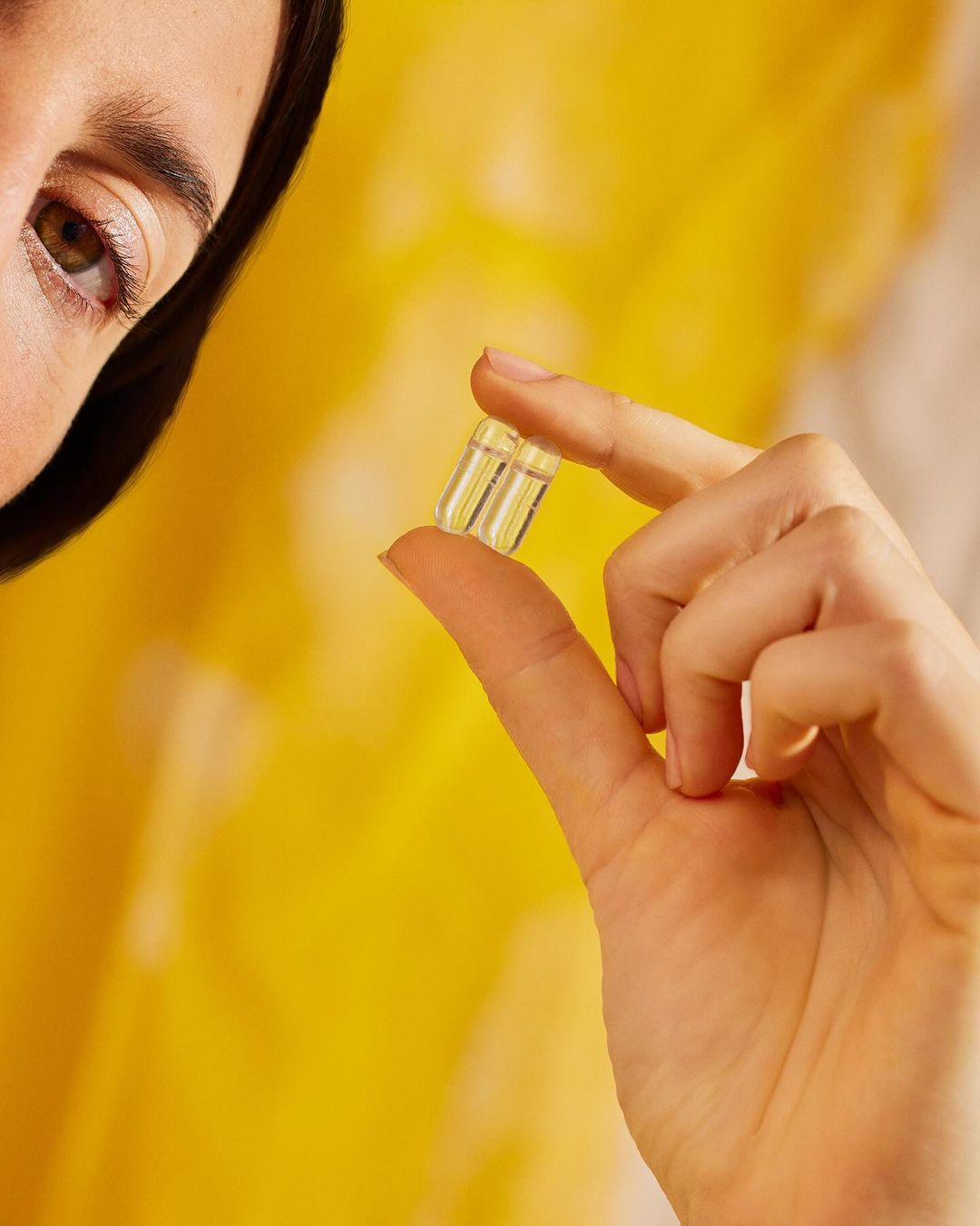 Close-up of an adult female holding two yellow gel capsules between her fingers, with a blurred yellow background.