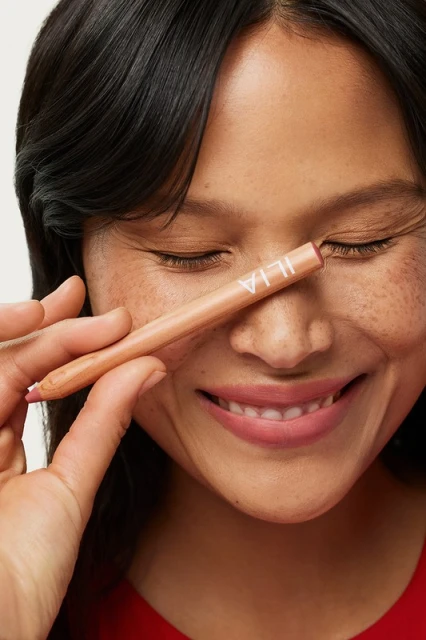 Close-up of a smiling woman with brown hair and freckles, posing with a lip pencil over her eye in a studio setting.