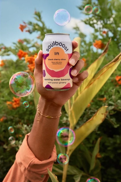 Hand holds a Soulboost lift black cherry citrus sparkling water can, surrounded by bubbles, with a leafy green and floral background.