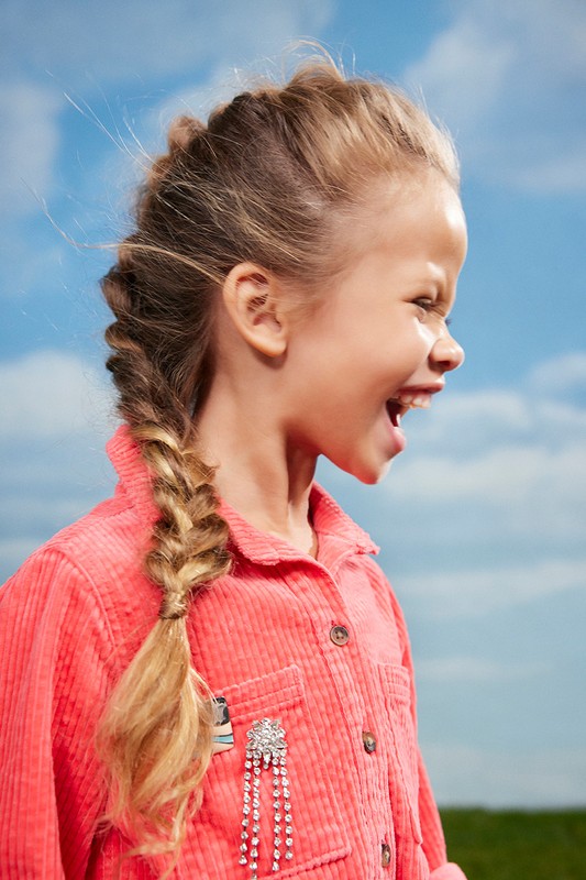 A young girl with a side braid and a vibrant coral shirt, standing against a blue sky, is captured in a medium outdoor shot.