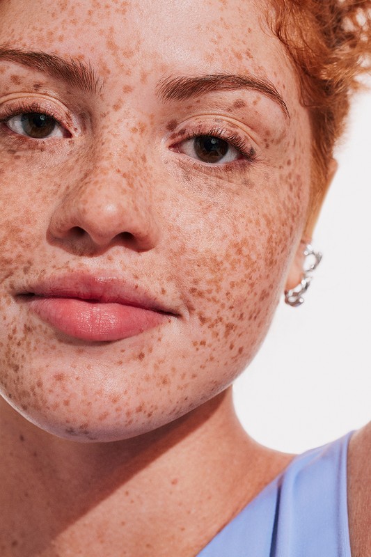 Close-up portrait of a woman with red hair and heavy freckles wearing a light blue top against a white background by Kat Borchart