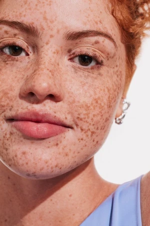 Close-up portrait of a woman with red hair and heavy freckles wearing a light blue top against a white background by Kat Borchart