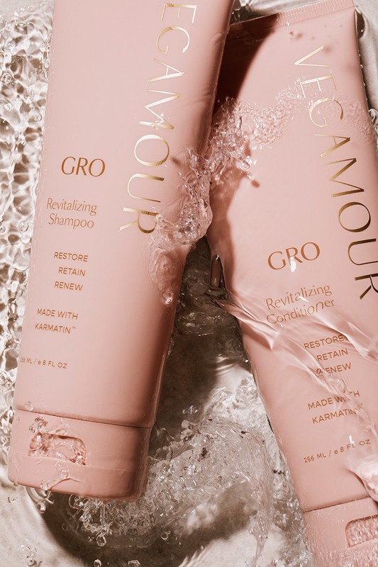 Two bottles of Vegamour GRO Revitalizing hair product, shampoo and conditioner, are photographed with water splashes in a soft, pinky beige light.