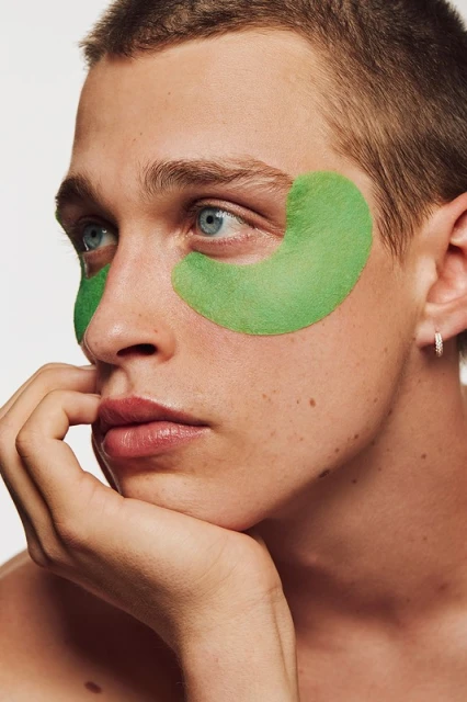 Close-up of a young man with green eye mask patches, fair skin, light eyes, and short hair, posing in a beauty or skincare context.