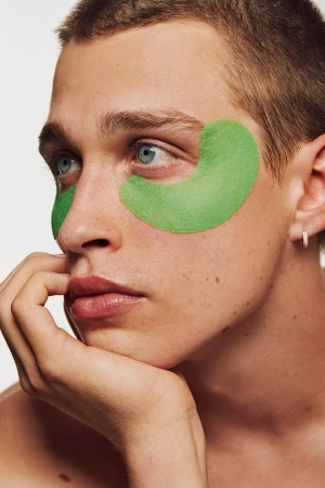 Close-up of a young man with green eye mask patches, fair skin, light eyes, and short hair, posing in a beauty or skincare context.