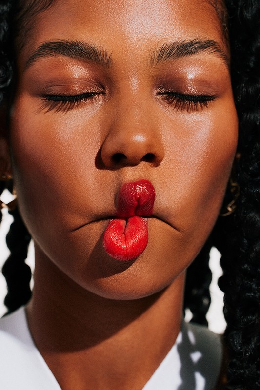 Close-up portrait of a woman with braided hair making a fish face with bright red lipstick against a plain white background.