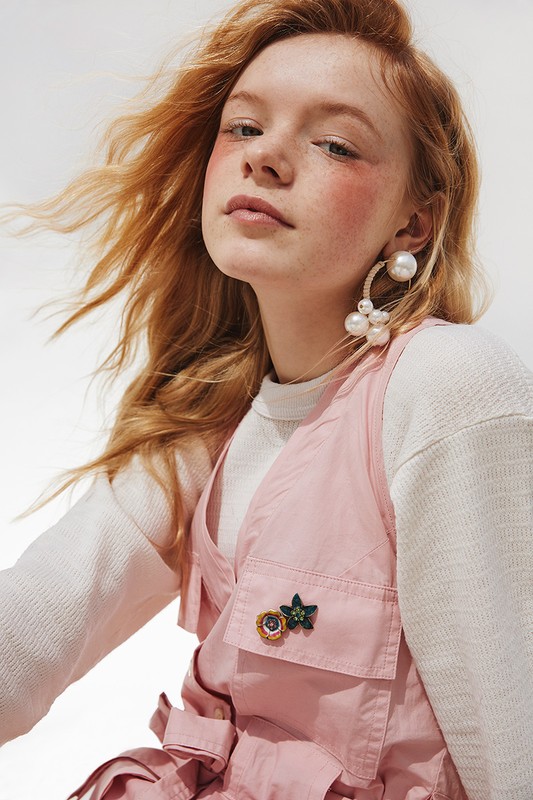 Portrait of a red-haired woman wearing pink vest and pearl earrings in a studio with bright, soft lighting, showcasing fashion and beauty.