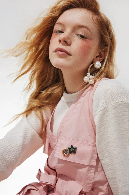 Portrait of a red-haired woman wearing pink vest and pearl earrings in a studio with bright, soft lighting, showcasing fashion and beauty.