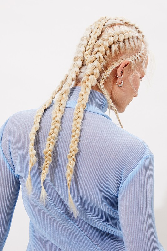 Back view of a fair-skinned adult female with blonde hair styled in cornrow braids, wearing a light blue ribbed turtleneck top in a studio.