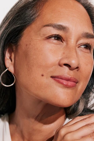 Close-up portrait of an older woman with silver-streaked dark hair, wearing a silver hoop earring and a white top.