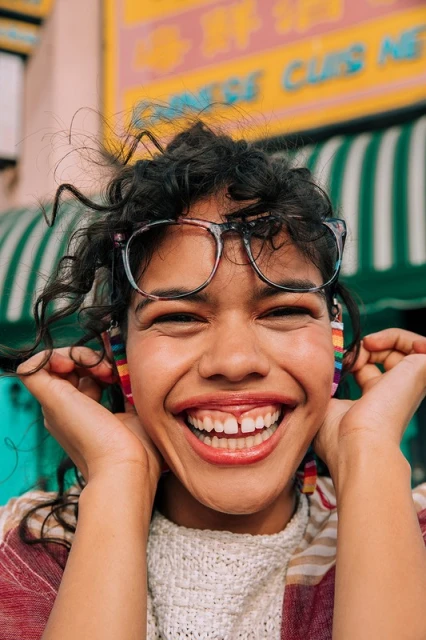 Close-up of a smiling young woman with glasses and curly hair, showcasing colorful earrings against a vibrant urban background.