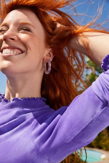 Close-up of a smiling red-haired woman outdoors wearing lilac earrings and a lilac top against a blue sky with palm trees.