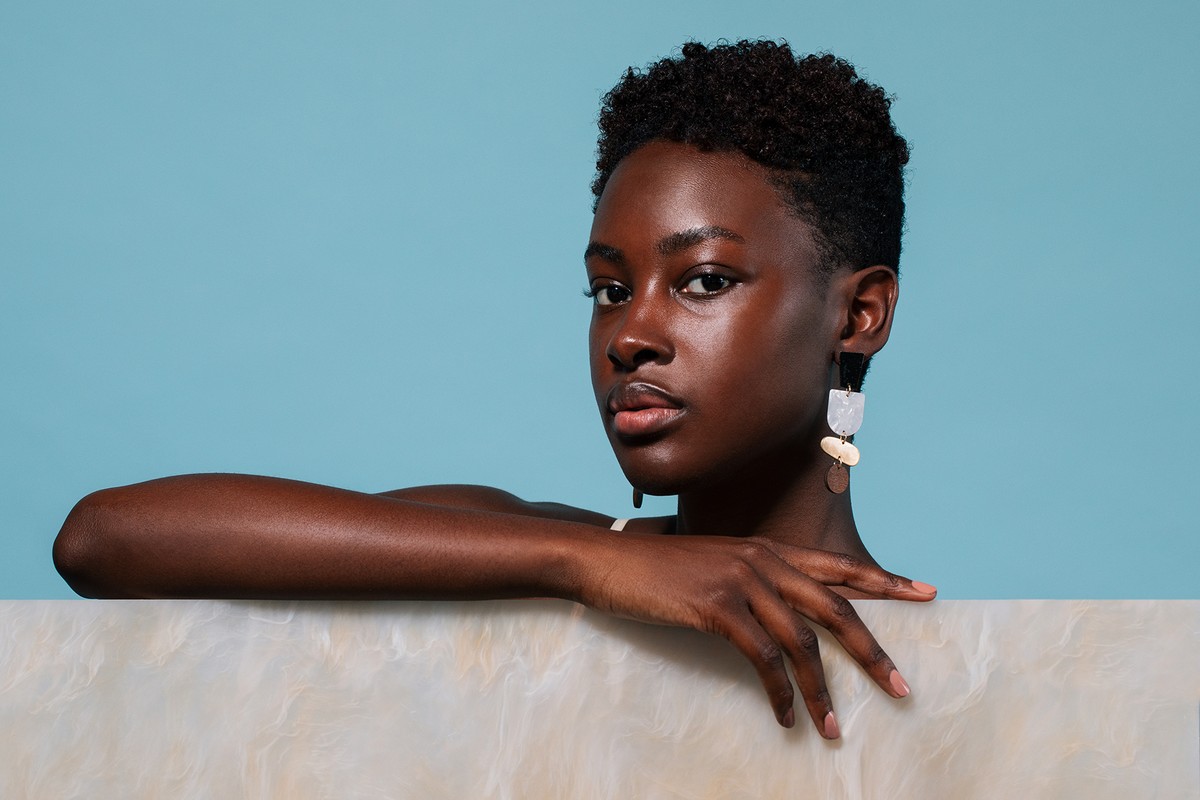 Close-up studio portrait of a young Black woman with curly hair, wearing modern earrings, against a light blue background.