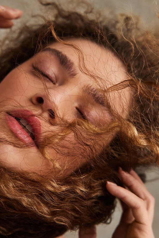 Close-up portrait of a woman with curly brown hair and closed eyes, with strands of hair falling across her face.