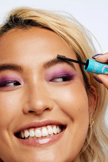 Close-up of an Asian woman applying brow gel with a spoolie brush, showcasing her vibrant purple eyeshadow and glossy lips in a beauty shot.