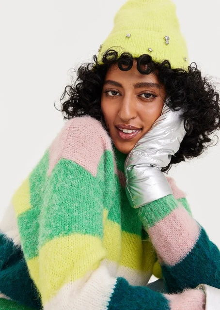 Attractive woman wearing a colorful striped sweater, neon yellow jeweled beanie and puffy silver glove, smiles in a studio shot.