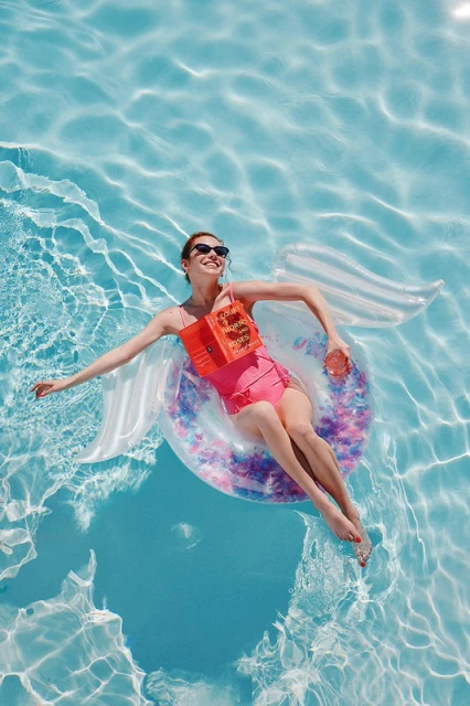 Woman floats in pool on inflatable ring reading book, wearing sunglasses and pink swimsuit in the bright sunlight of a summer day.