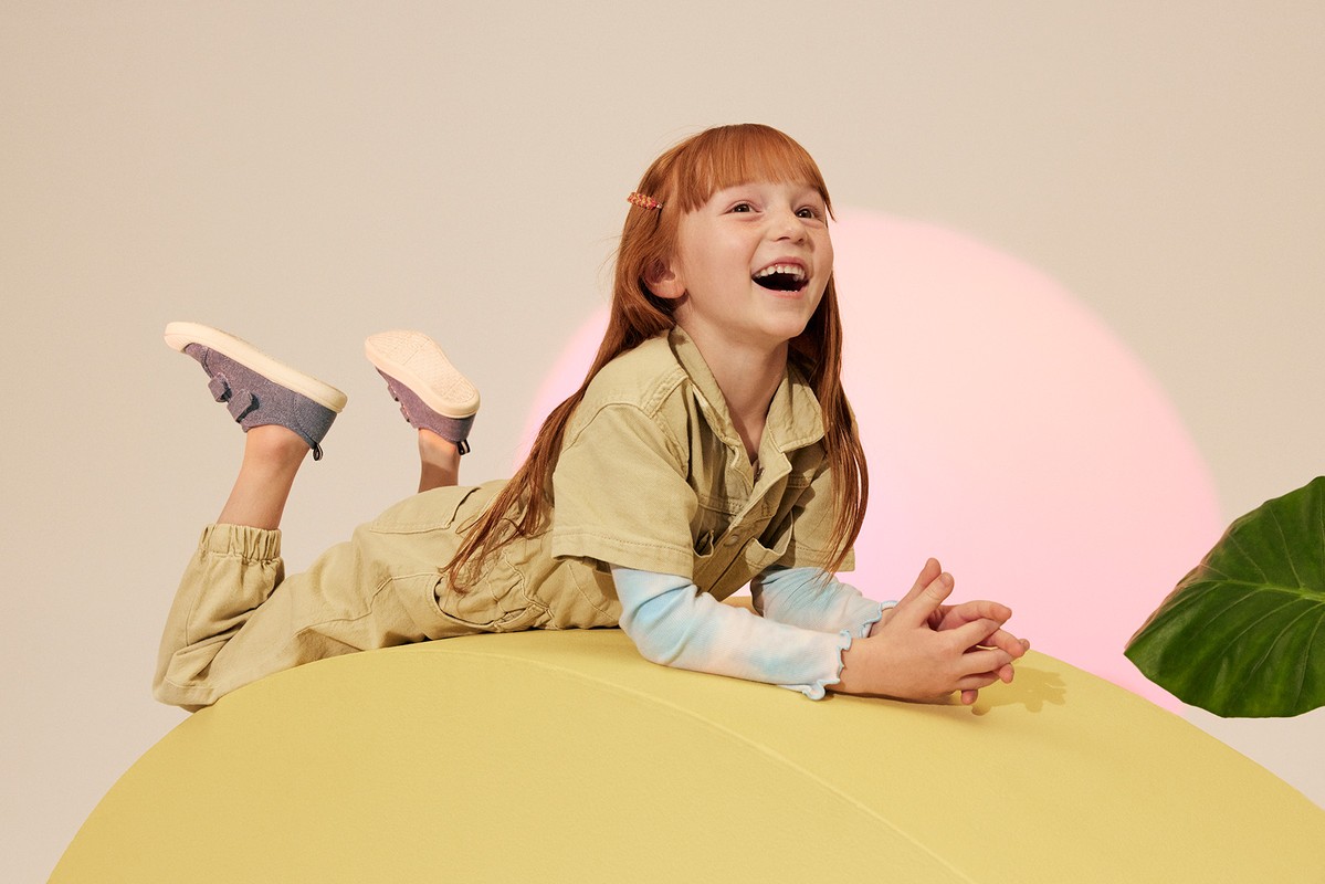 Red-haired child in a khaki jumpsuit laughs while lying on a large yellow ball in a studio setting with a pink spotlight.