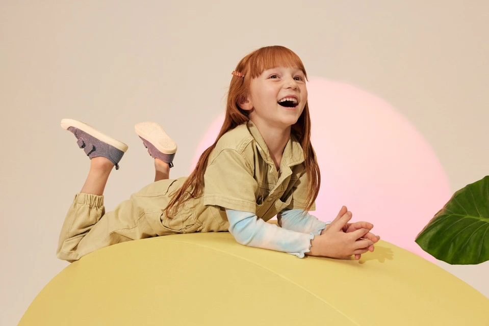 Red-haired child in a khaki jumpsuit laughs while lying on a large yellow ball in a studio setting with a pink spotlight.