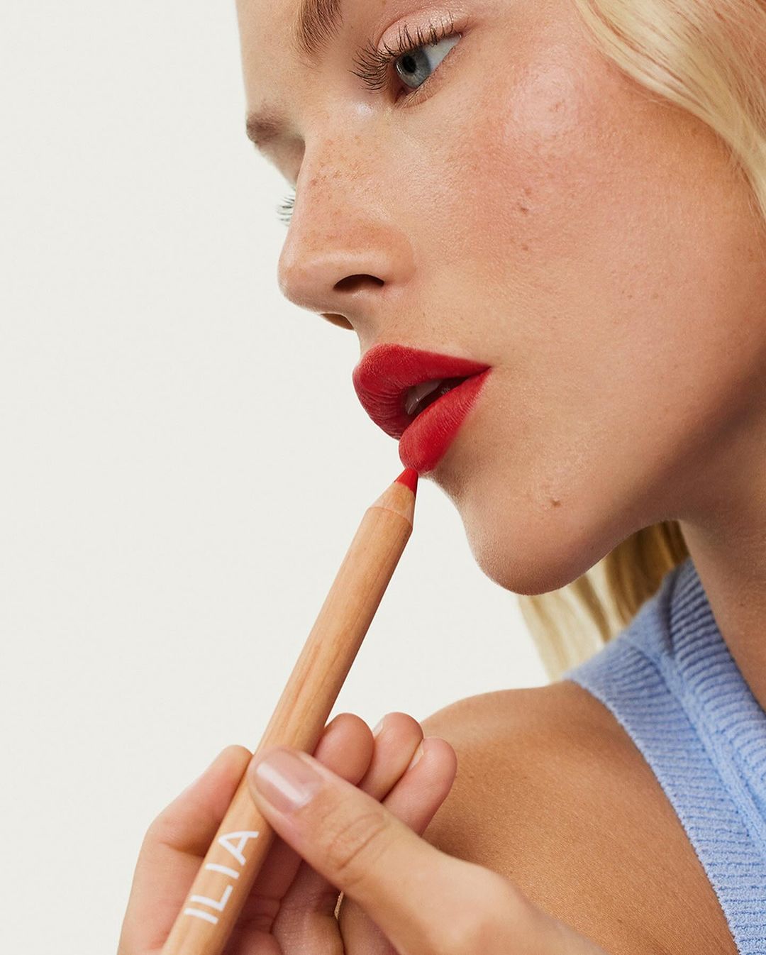 Close-up of a fair-skinned adult woman applying red lip liner. Makeup focuses on the lips with a bright red color against a white background.