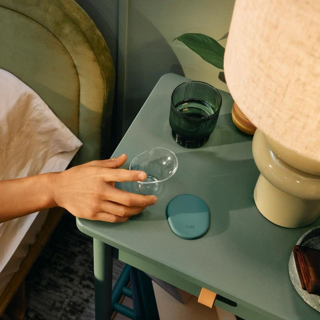 Close-up of a hand reaching for an empty glass on a green table next to another glass, a lamp, and a bed in the background.