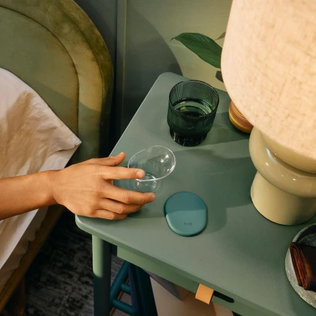 Close-up of a hand reaching for an empty glass on a green table next to another glass, a lamp, and a bed in the background.
