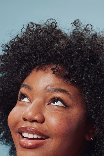 Close-up portrait of a smiling Black woman with curly hair looking up against a light blue backdrop in a beauty or fashion context.