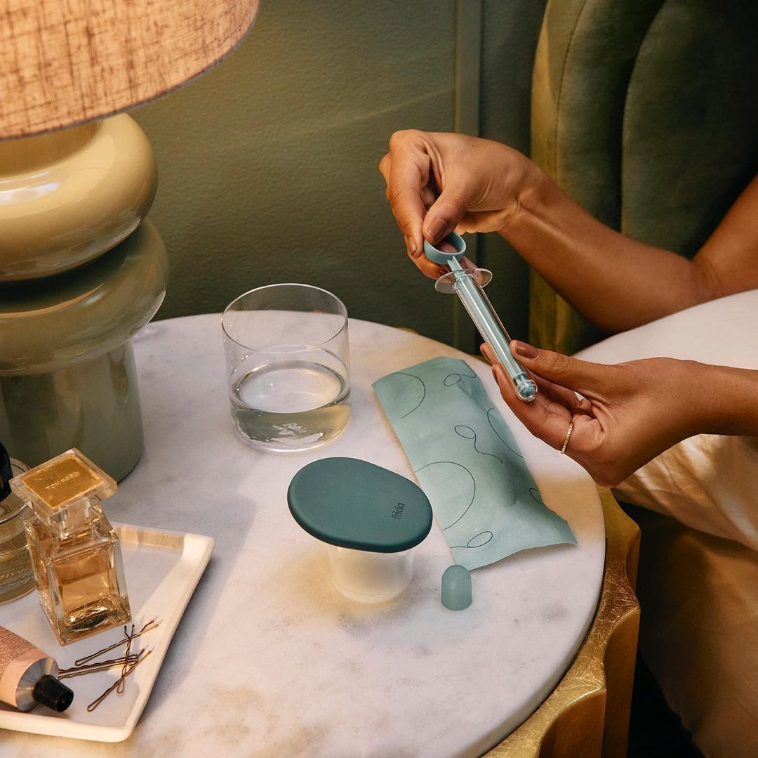 Close-up of a syringe held by woman's hands on a marble table with perfume bottles, lamp, and a glass of water in soft lighting.