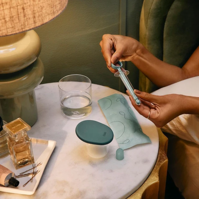 Close-up of a syringe held by woman's hands on a marble table with perfume bottles, lamp, and a glass of water in soft lighting.
