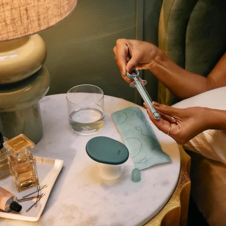 Close-up of a syringe held by woman's hands on a marble table with perfume bottles, lamp, and a glass of water in soft lighting.